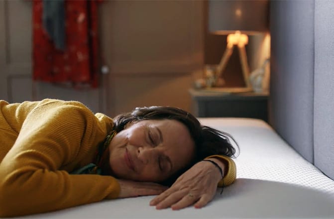 Persona distesa su un materasso con maglone giallo, lampada e vestiti sullo sfondo.