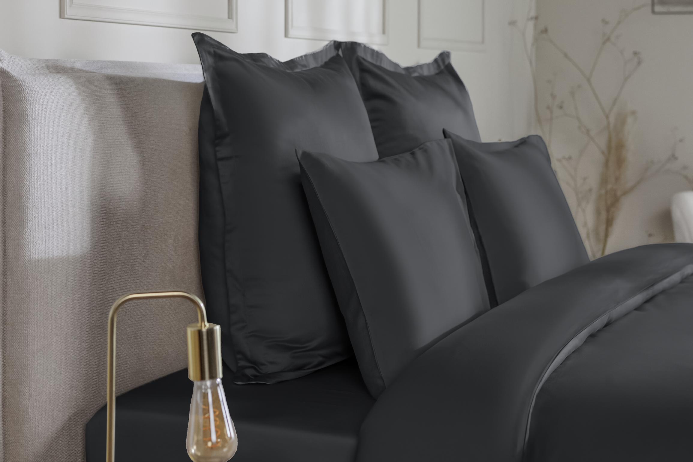 Zes donkergrijze Emma Satijnen Kussenslopen gepresenteerd op een bed: drie grote kussens achteraan en drie kleinere ervoor, allemaal voorzien van gladde, glanzende satijnstof in een diepe grijstint; bed is opgemaakt met bijpassend donkergrijs dekbed; achtergrond toont een lichtgekleurde wand met sierpanelen, rechts een moderne lamp en een vaas met droogbloemen.