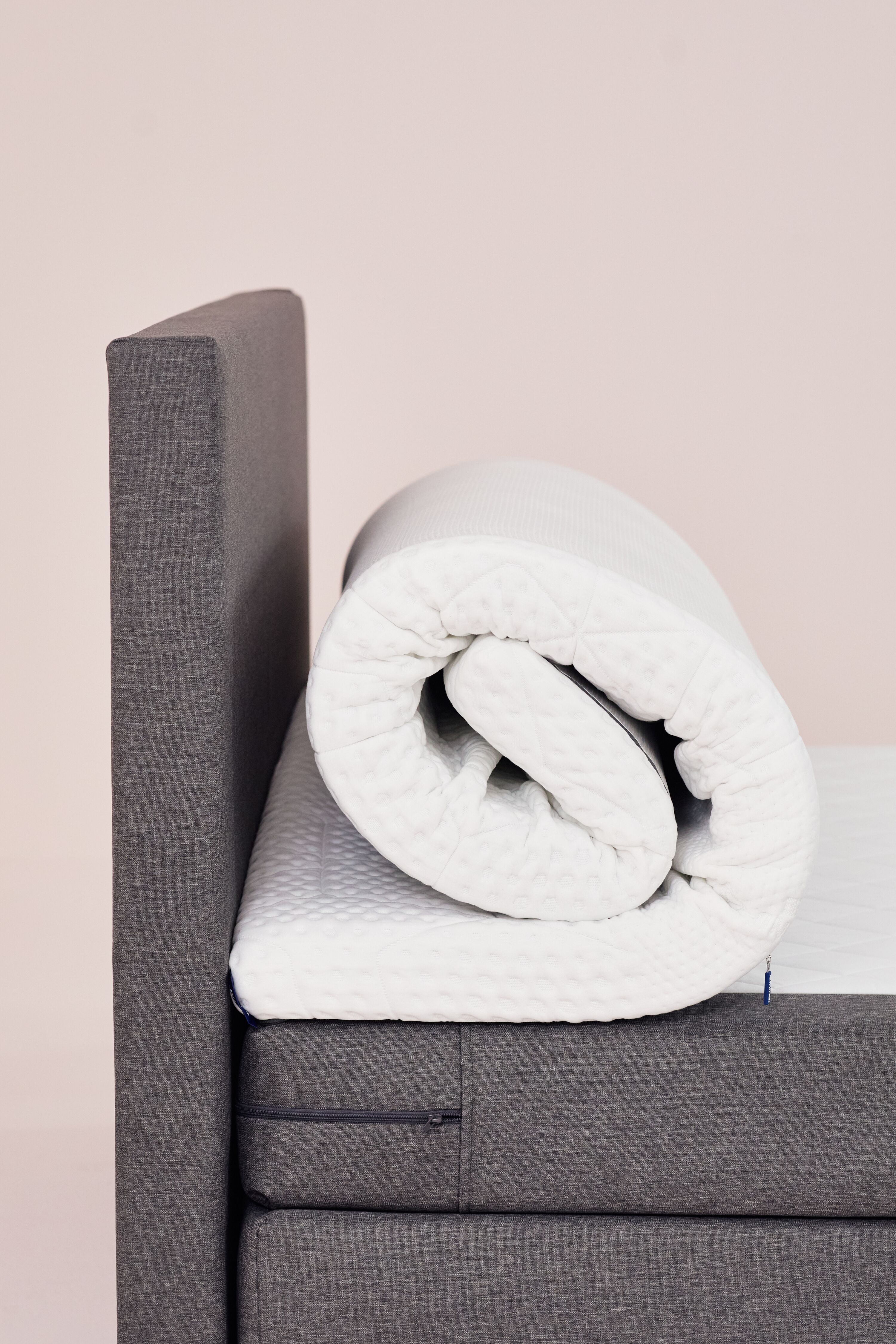 Aufgerollter Emma Topper auf einem Bett mit grauem Kopfteil.