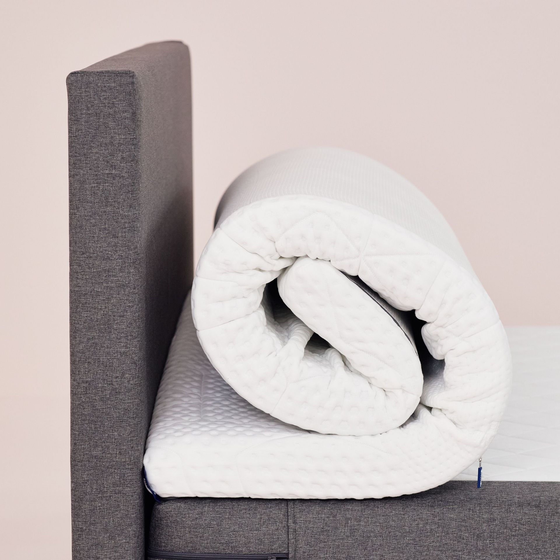 Aufgerollter Emma Topper auf einem Bett mit grauem Kopfteil.