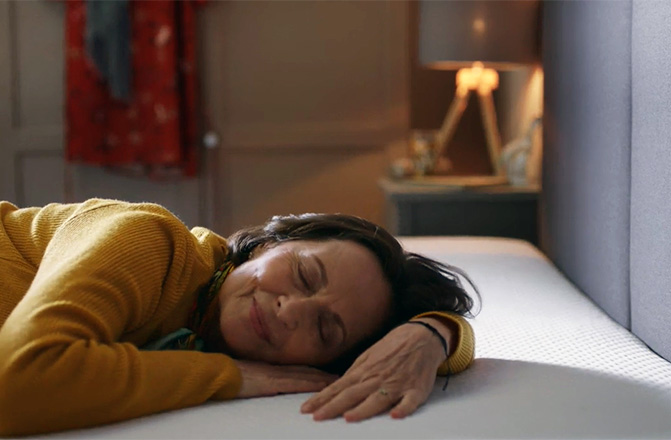Personne allongée sur un matelas, souriante et détendue, dans une chambre chaleureuse avec lampe allumée et vêtements suspendus en arrière-plan.