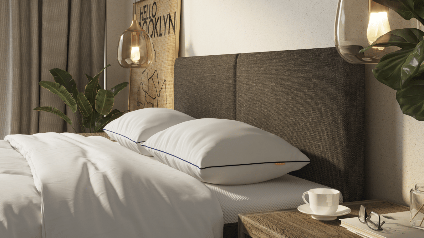 Bed met witte lakens en twee witte kussens met blauwe biezen; donker hoofdbord, nachtkastje met kopje, bril en boek; glazen hanglamp en wanddecoratie.