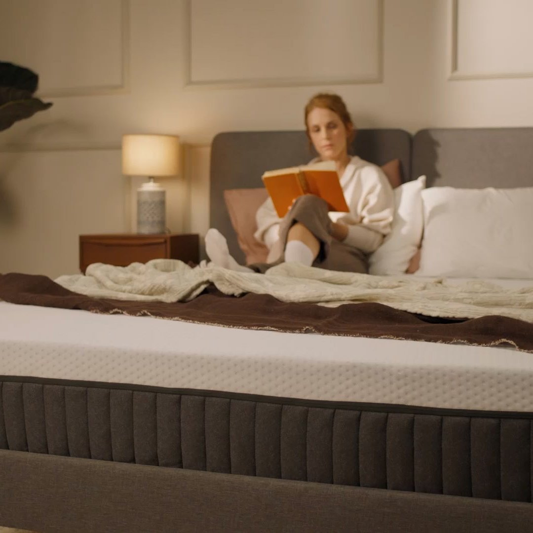 Person sitting on a bed with a white mattress in a bright bedroom.