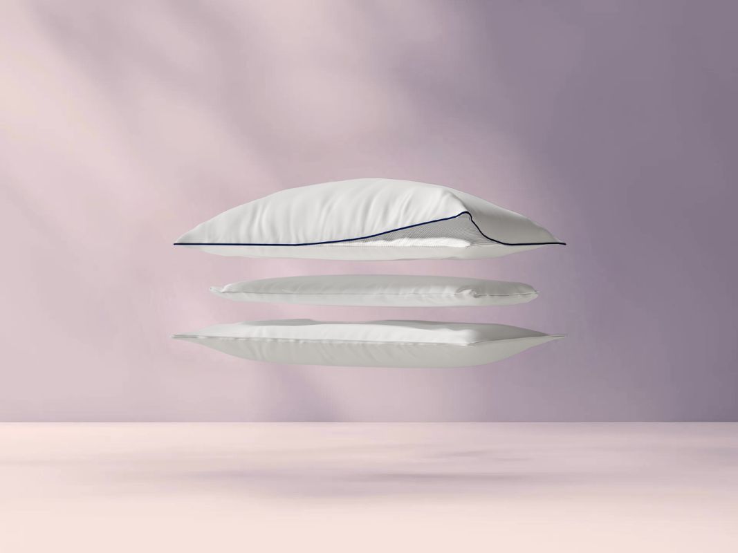 Drie gestapelde lagen van het Emma Premium Wolkenkussen zweven tegen pastelroze achtergrond; bovenste laag omgevouwen toont rasterpatroon van binnenlaag, blauwe biezen zichtbaar.