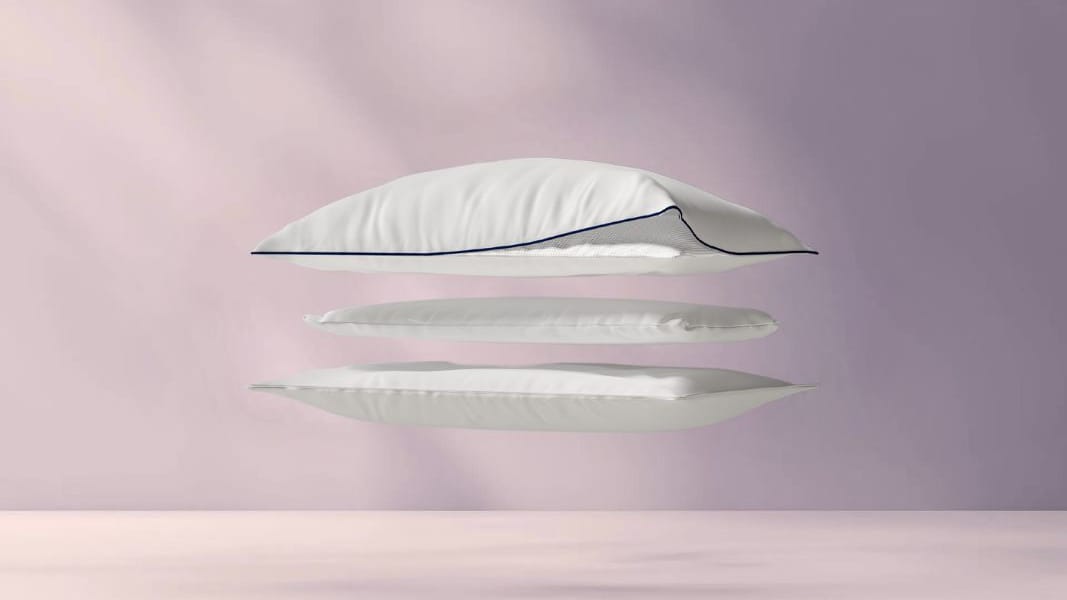 Drie gestapelde lagen van het Original Cooling Comfort Kussen zweven tegen pastelroze achtergrond; bovenste laag omgevouwen toont rasterpatroon van binnenlaag, blauwe biezen zichtbaar.