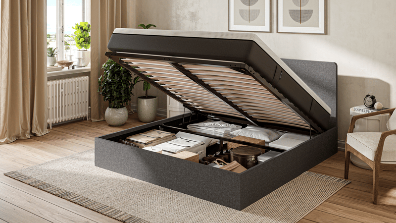 Une chambre à coucher moderne avec un lit de rangement relevable révélant des objets bien rangés sous le lit, notamment des boîtes, des oreillers et des couvertures. La lumière du soleil passe à travers une fenêtre aux rideaux beiges, mettant en valeur un décor confortable et neutre.