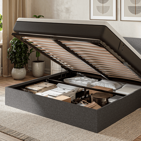 Une chambre à coucher moderne avec un lit de rangement relevable révélant des objets bien rangés sous le lit, notamment des boîtes, des oreillers et des couvertures. La lumière du soleil passe à travers une fenêtre aux rideaux beiges, mettant en valeur un décor confortable et neutre.