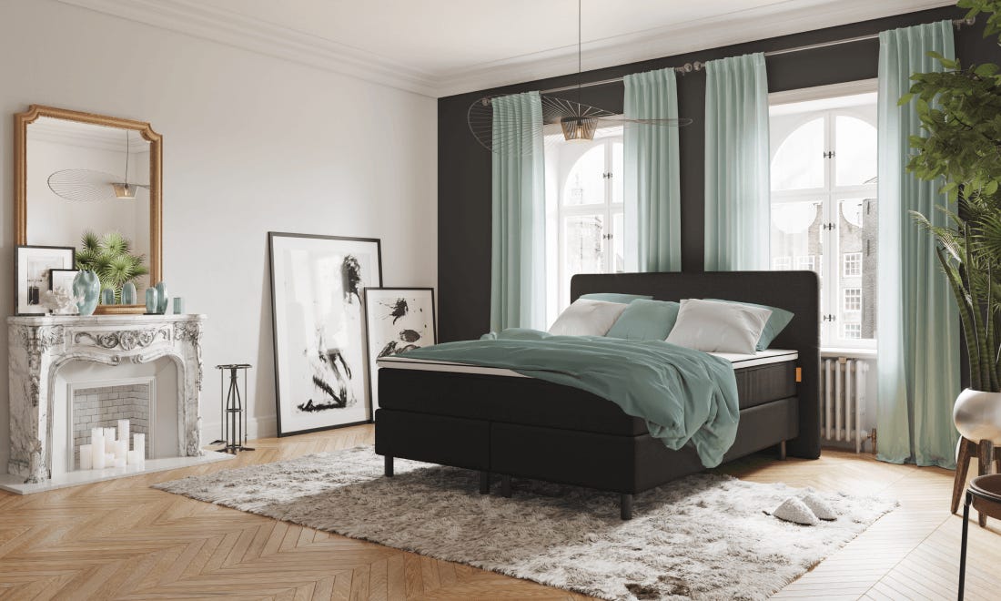 Emma Deluxe Boxspring met zwart frame, teal beddengoed en witte kussens in elegante slaapkamer met grote spiegel, open haard, houten vloer en decoratieve accenten.
