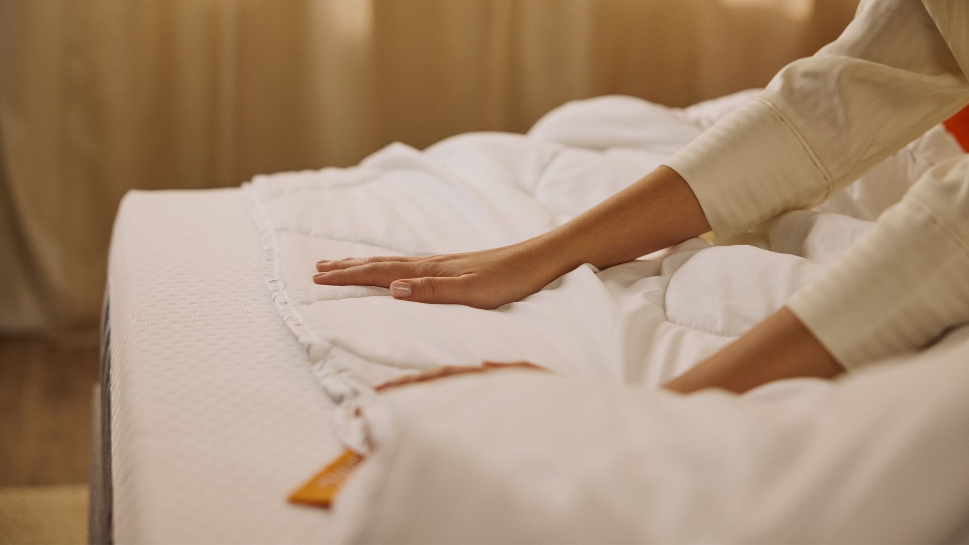 Handen die een Emma dekbed gladstrijken op een bed, met zachte witte stof en een label zichtbaar aan de rand.