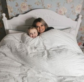 Mum and son lying in bed under a white blanket, with a carved white headboard and floral-patterned wallpaper in the background.