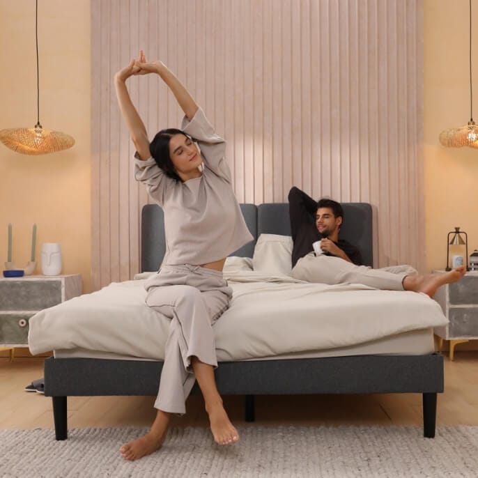 Deux personnes sur un lit dans une chambre moderne : l’une assise au bord du lit les bras levés, l’autre adossée à la tête de lit tenant une tasse. Lit avec cadre foncé et linge clair, éclairé par deux suspensions tressées et entouré de tables de chevet décorées.