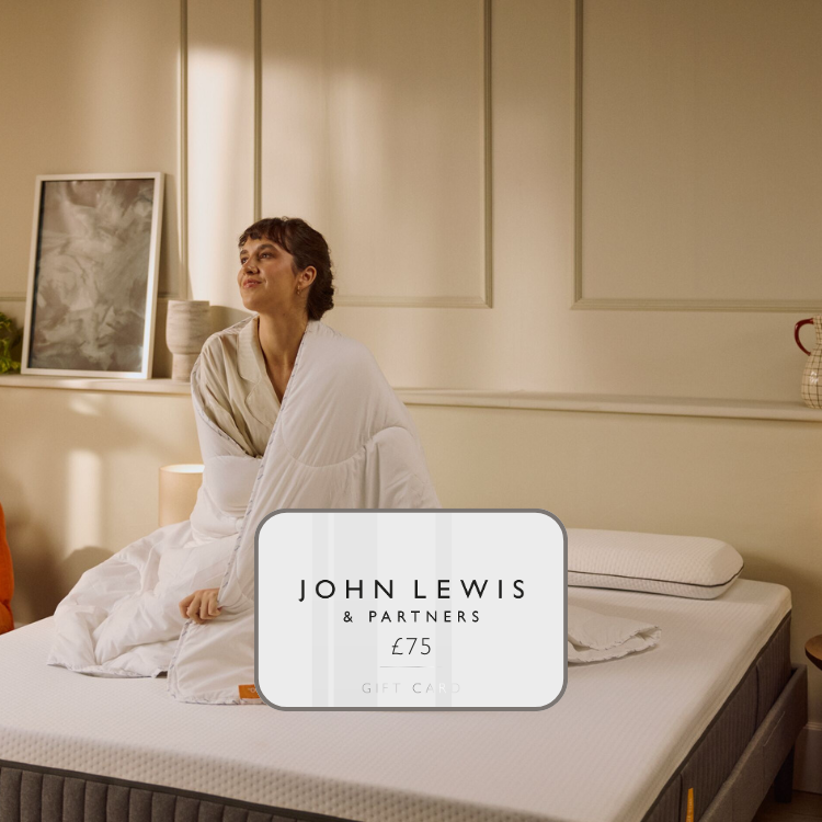 A woman wrapped in a white duvet, sitting on top of an Emma Mattress in a well light bedroom.