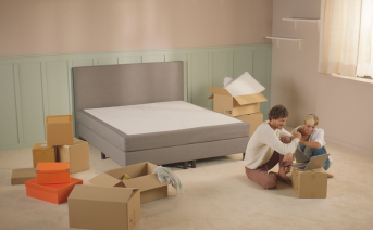 A couple crouched next to a closed Emma box in a room full with boxes and an assembled bed. The couple is looking at a computer.