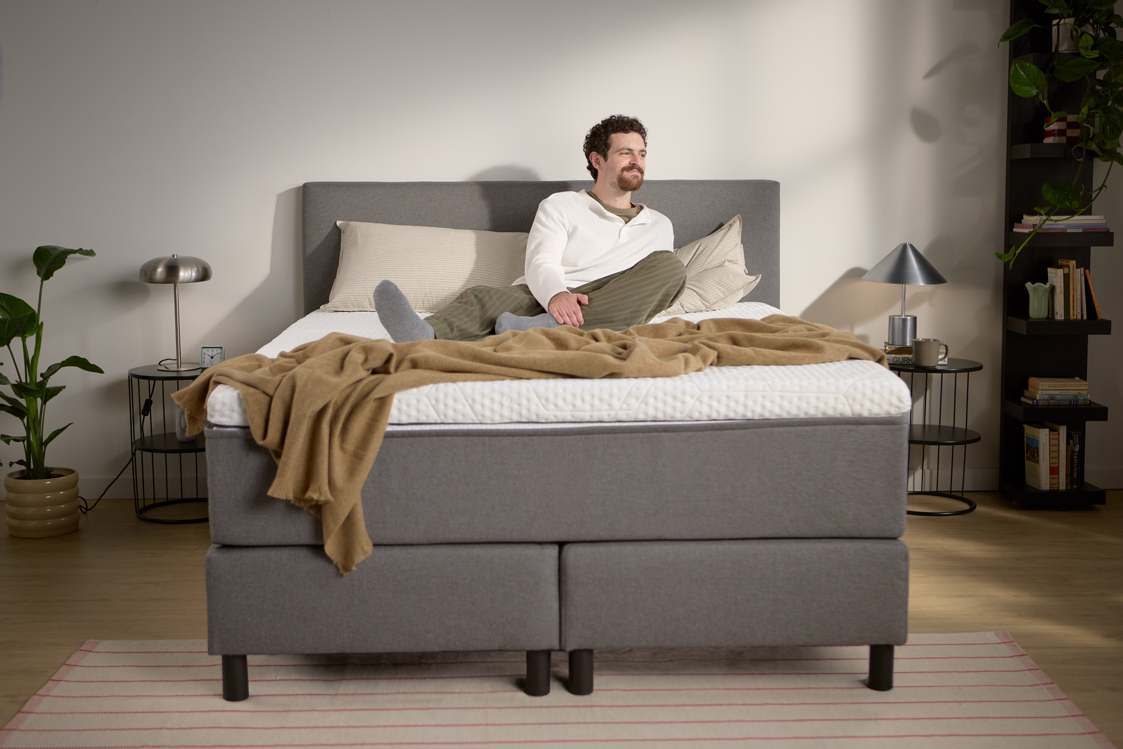 Lifestyle beeld met een persoon zittend op het grijze boxspringbed; toont de hoogte van matras en topper, kussens en een lichte plaid.