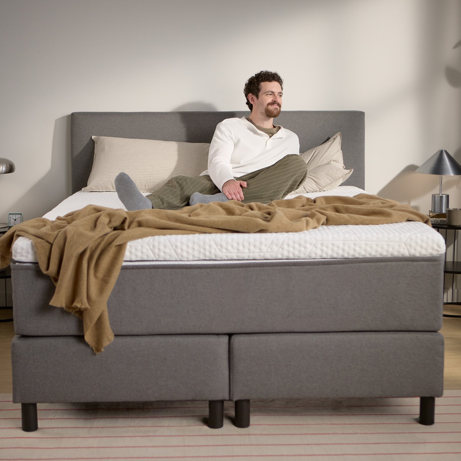 Lifestyle beeld met een persoon zittend op het grijze boxspringbed; toont de hoogte van matras en topper, kussens en een lichte plaid.