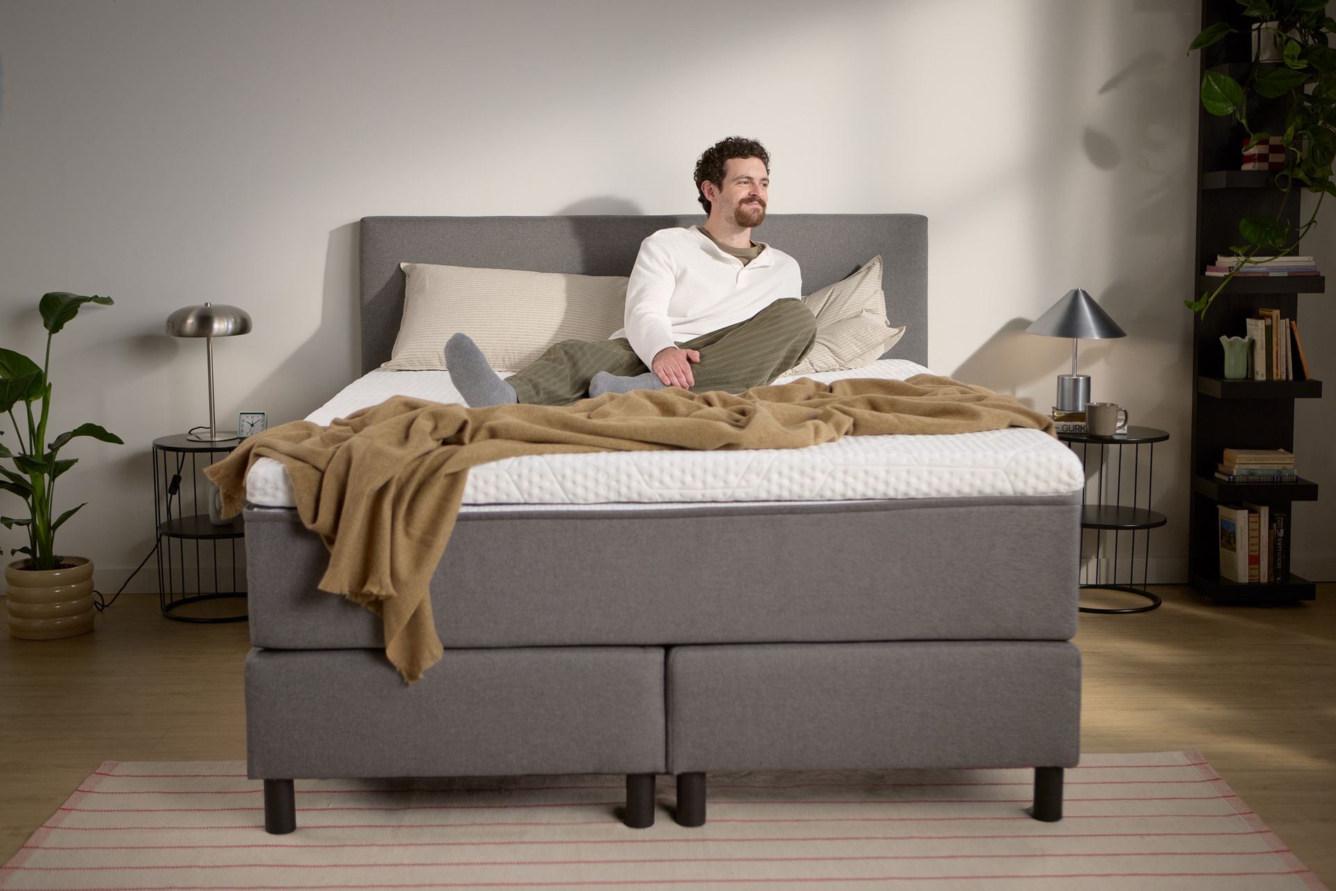 Lifestyle beeld met een persoon zittend op het grijze boxspringbed; toont de hoogte van matras en topper, kussens en een lichte plaid.