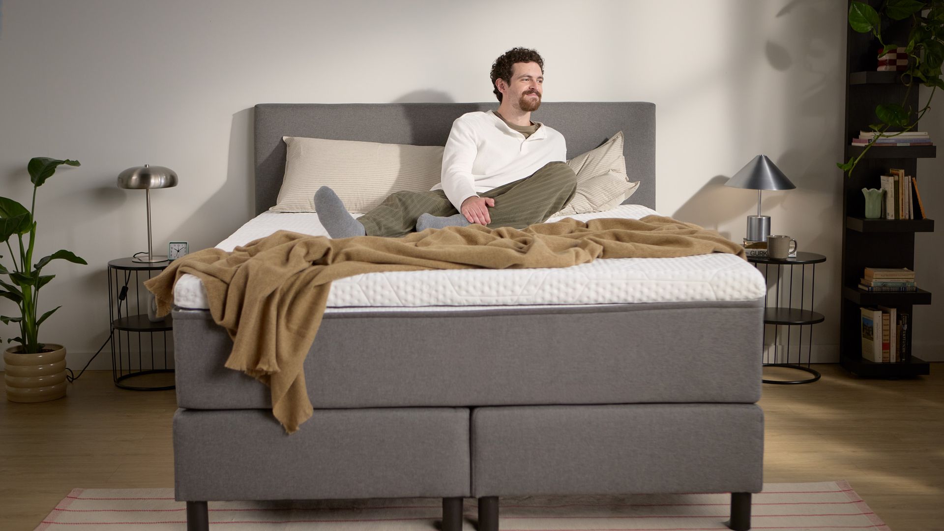 Lifestyle beeld met een persoon zittend op het grijze boxspringbed; toont de hoogte van matras en topper, kussens en een lichte plaid.