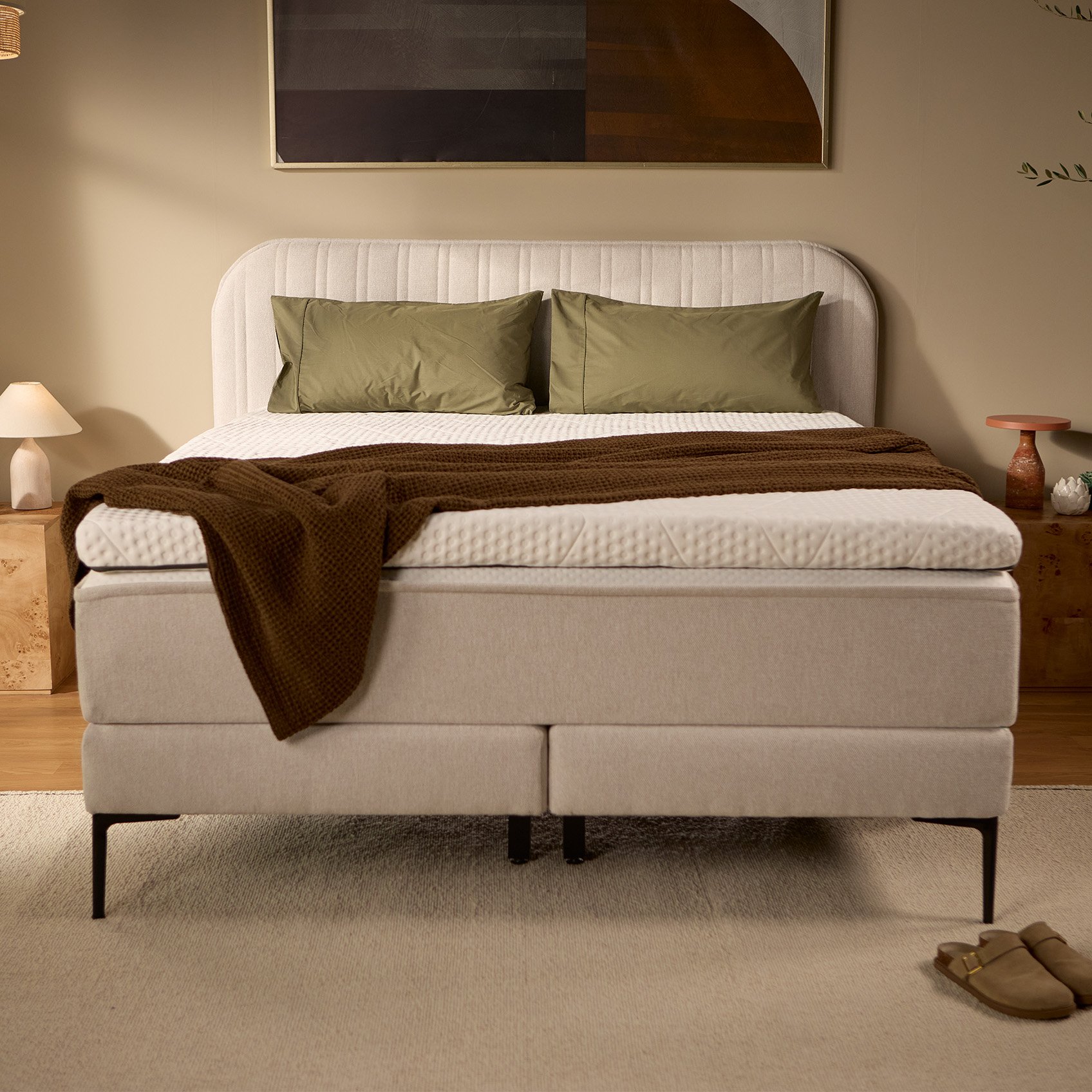 Light beige upholstered box spring bed with channel-stitched headboard, pillows, and bedding, shown from the front.