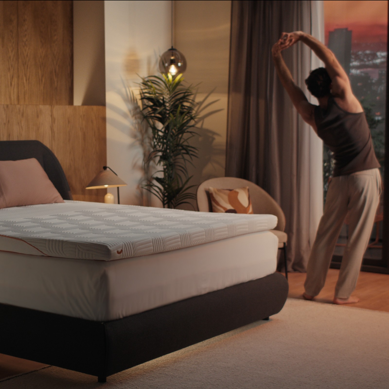 Man stretching by bedroom window at sunrise next to dark boxspringbed with Emma Performance Topper.