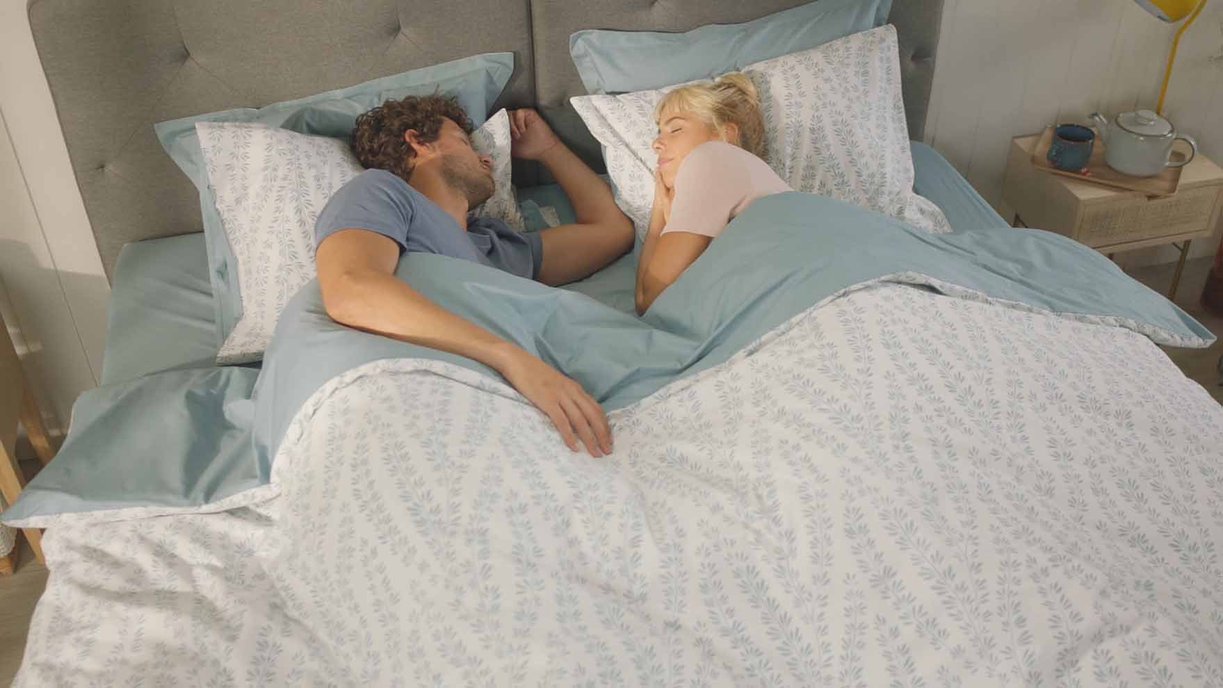 Deux personnes allongées face à face dans un lit, sous une couette bleu clair à motifs, dans une chambre paisible avec éclairage doux et table de nuit garnie d’objets du quotidien.