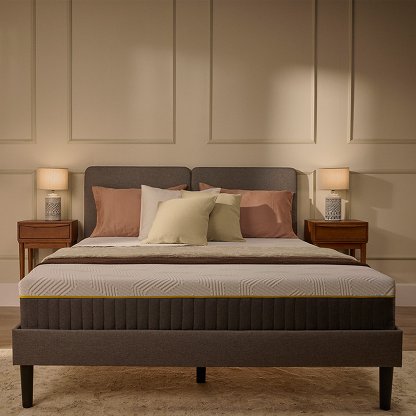 Emma Original Pro mattress with white textured cover and yellow trim, styled on a dark grey bed frame in a warm bedroom.
