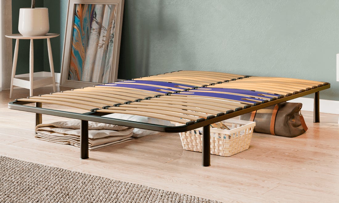 Emma Essential Bedframe met houten lattenbodem en zwart metalen frame op houten vloer—groene muur, abstract schilderij, opbergmand en dekens onder het bed benadrukken functionaliteit en minimalistisch design.