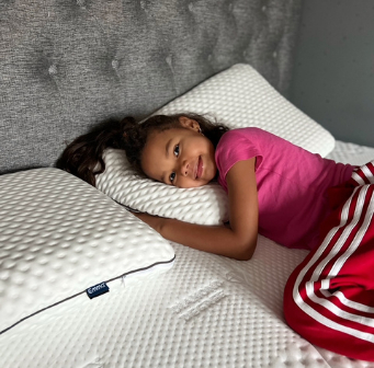 Kind ligt ontspannen op een bed met de Emma Diamond Topper, omringd door witte kussens en een gestoffeerd grijs hoofdbord—een topper met diamantstructuur die zacht en temperatuurregulerend comfort biedt.