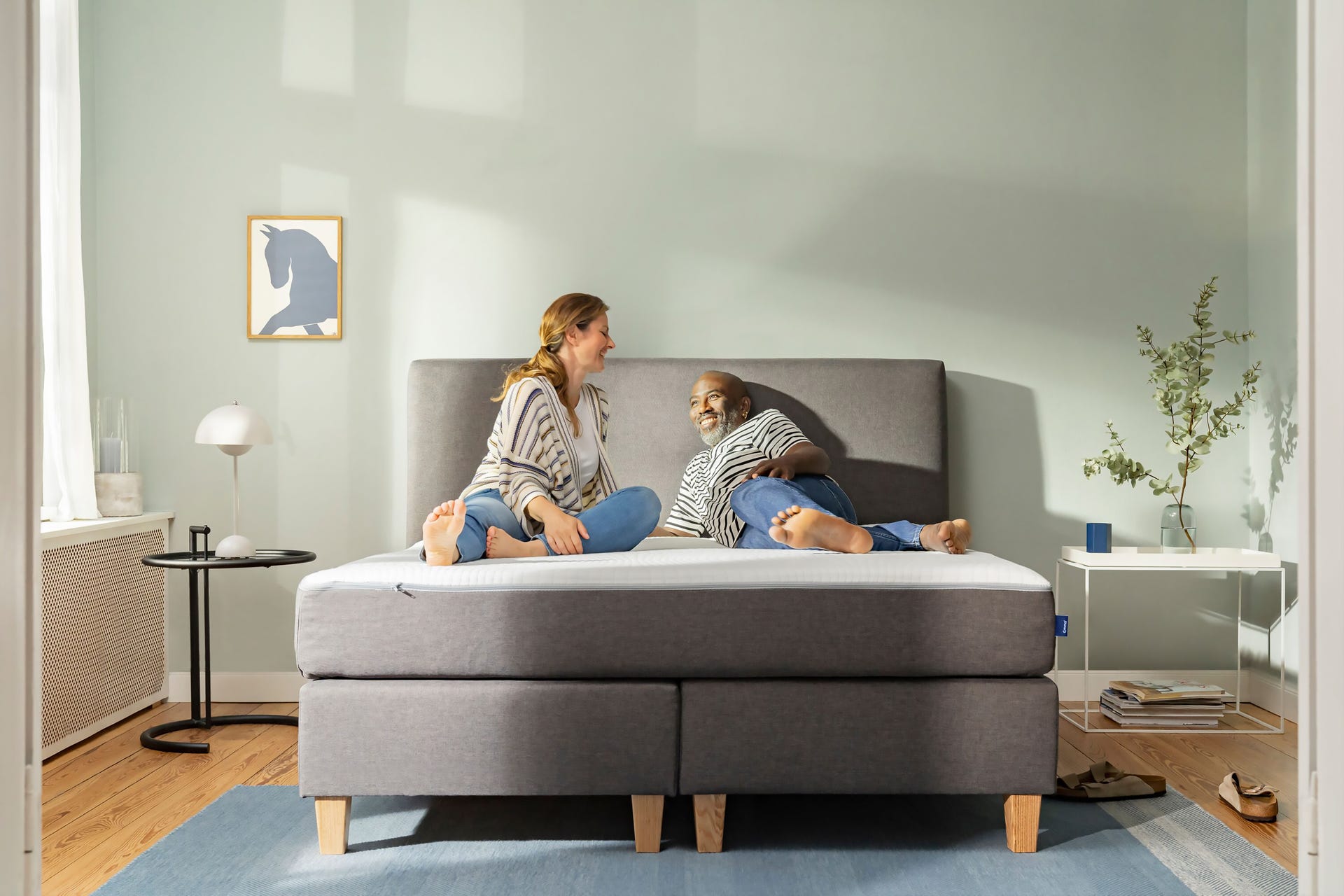 Twee personen zitten op een moderne, grijze Emma Deluxe Boxspring in een minimalistische slaapkamer met houten vloer, lichtgekleurde muren en natuurlijk licht. Naast het bed staat een wit nachtkastje met een lamp en decoratie; aan de muur hangt een kunstwerk van een blauwe hond.