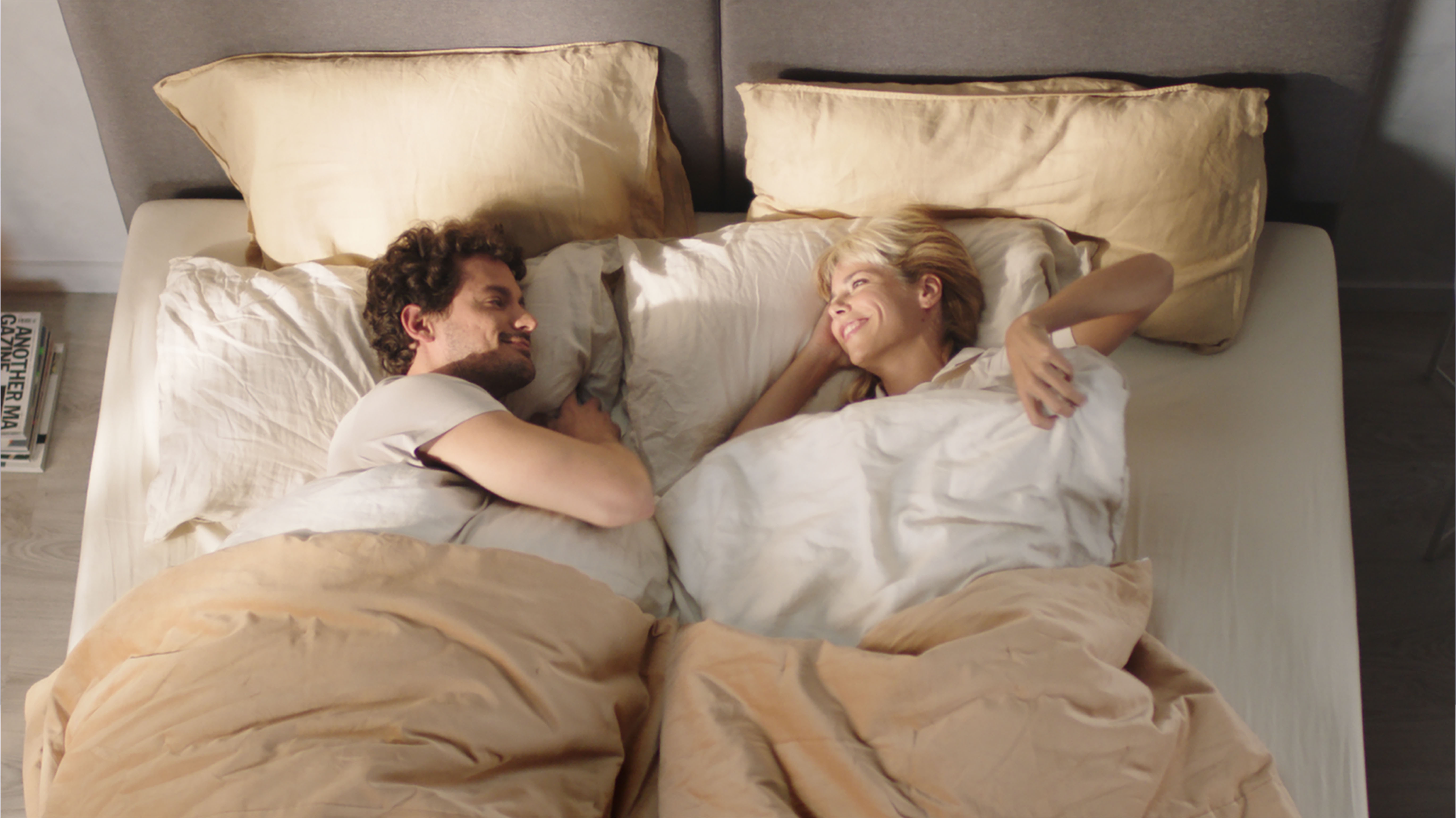 Deux personnes allongées face à face dans un lit, souriantes et détendues sous une literie beige et blanche. La scène évoque un moment intime et chaleureux, avec deux oreillers visibles et un magazine posé au sol près du lit.