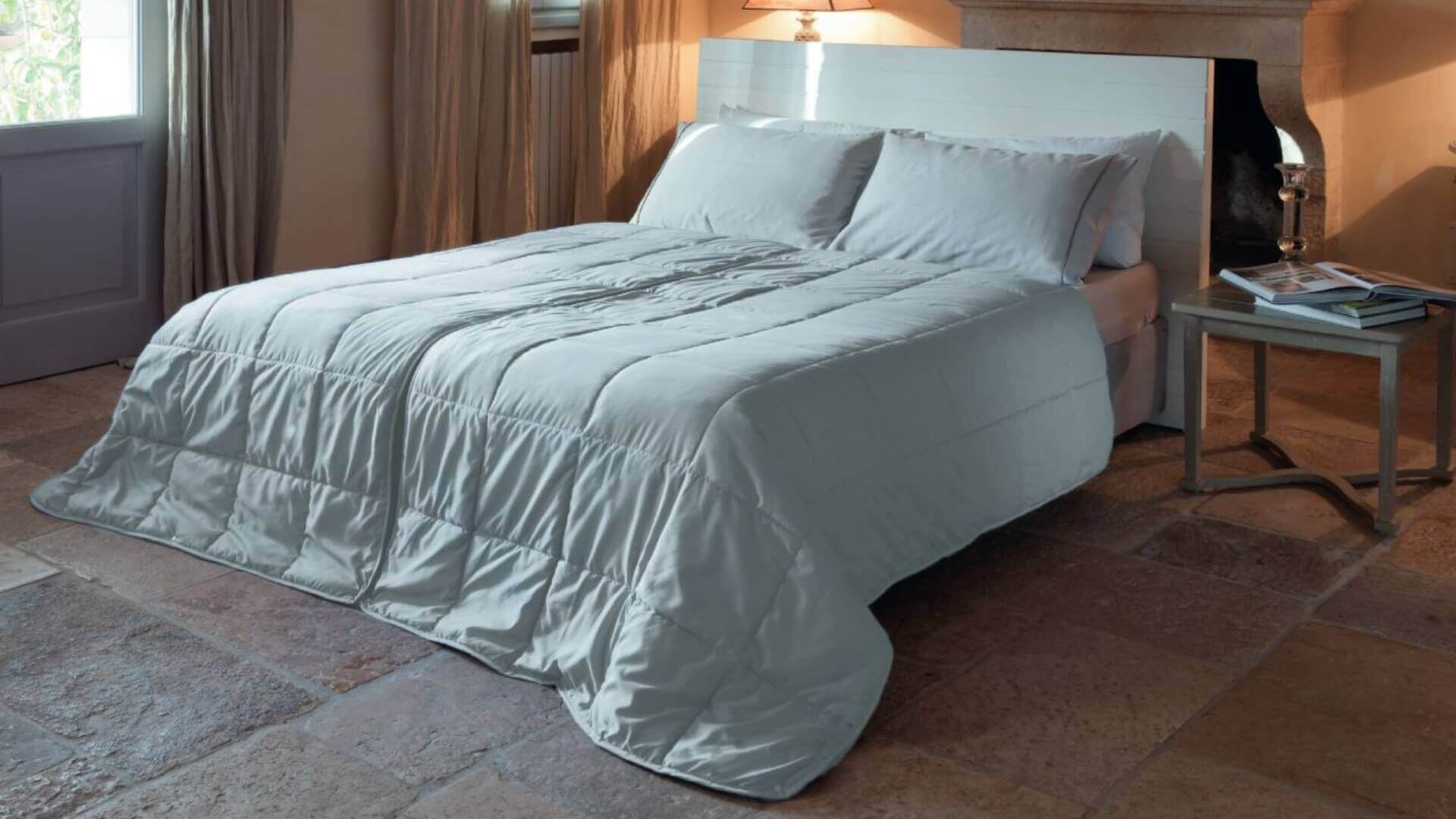 Letto con biancheria bianca in camera accogliente, con comodino, lampada, camino e luce naturale.