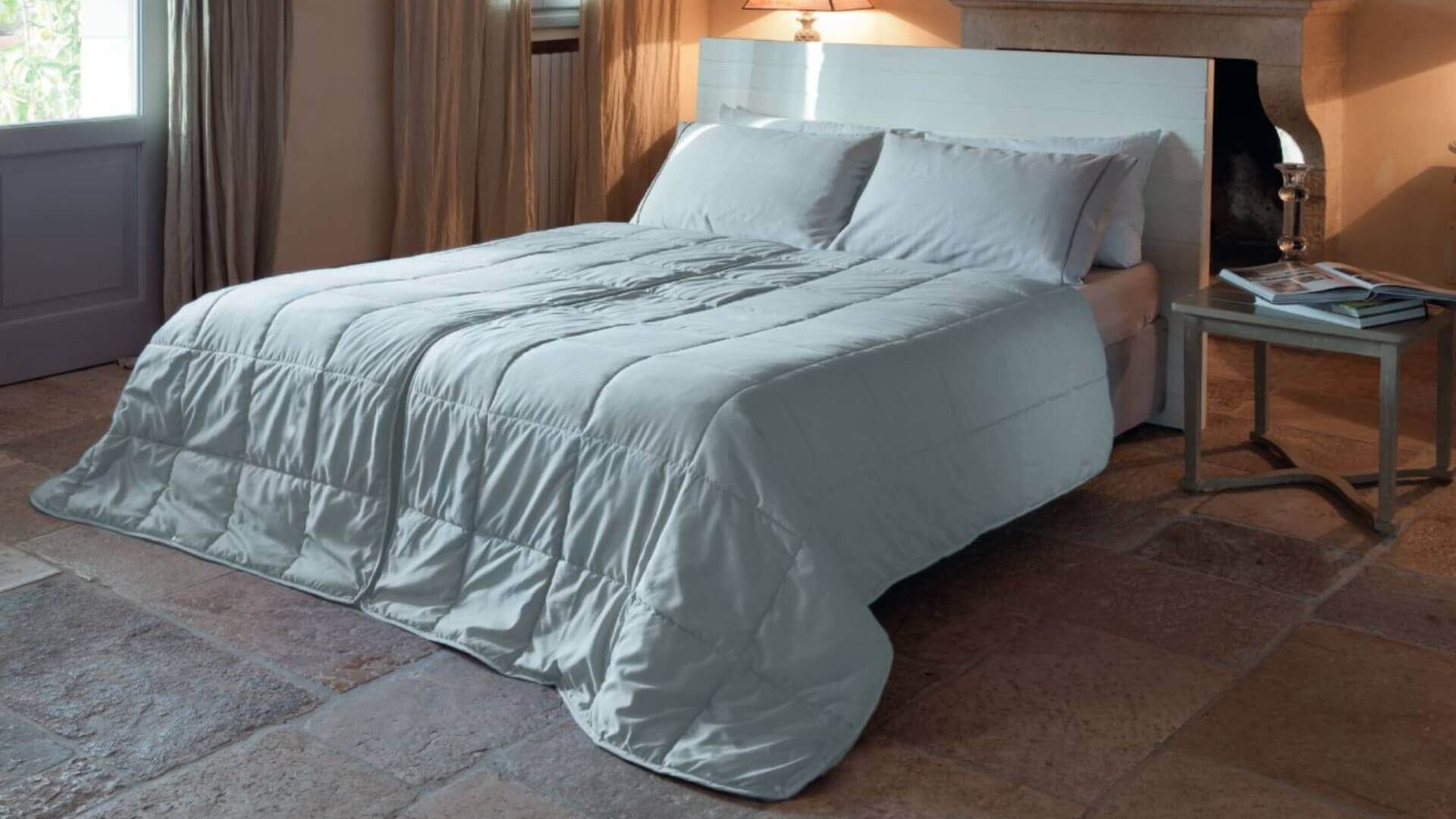 Letto con biancheria bianca in camera accogliente, con comodino, lampada, camino e luce naturale.