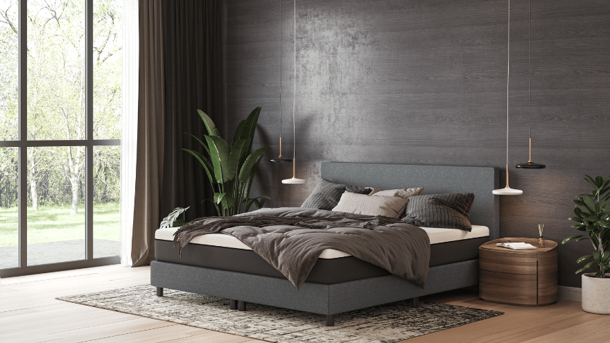 Lit avec tête de lit et base rembourrées en gris, habillé de linge de lit gris foncé et blanc, avec plusieurs oreillers. Placé sur un tapis à motifs sur parquet clair. Deux tables de chevet rondes en bois de chaque côté, chacune surmontée d’une suspension. Mur arrière en panneaux de bois foncé. À gauche, grande fenêtre avec rideaux clairs et foncés, vue sur des arbres. Plante verte près de la fenêtre. Ambiance minimaliste, élégante et lumineuse.