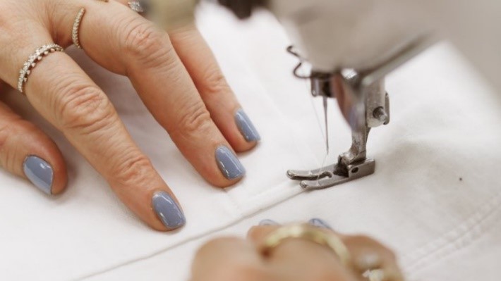 Nahaufnahme einer Nähmaschine beim Nähen von weißem Stoff, geführt von Händen mit blau lackierten Fingernägeln.