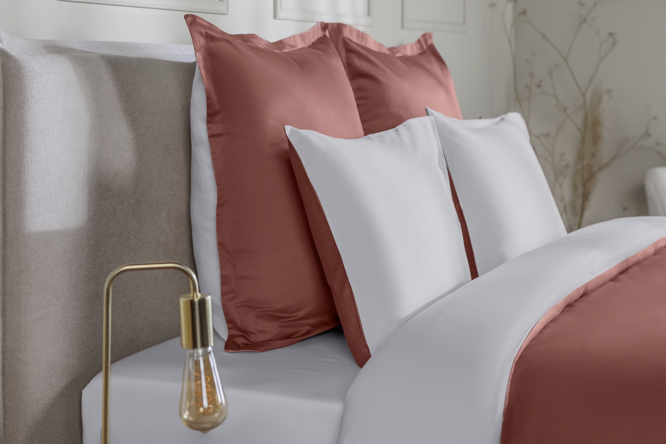 Detail van een bedhoek met een Emma Satijn Hoeslaken in een warme terracottakleur, strak om een matras gespannen; hoeslaken heeft een subtiele glans en zijdezachte afwerking die het rijke aardetint benadrukt; bedframe is grijs met houten poten; setting toont een witte muur met sierpanelen, links een lamp met gouden voet en rechts een vaas met droogbloemen.