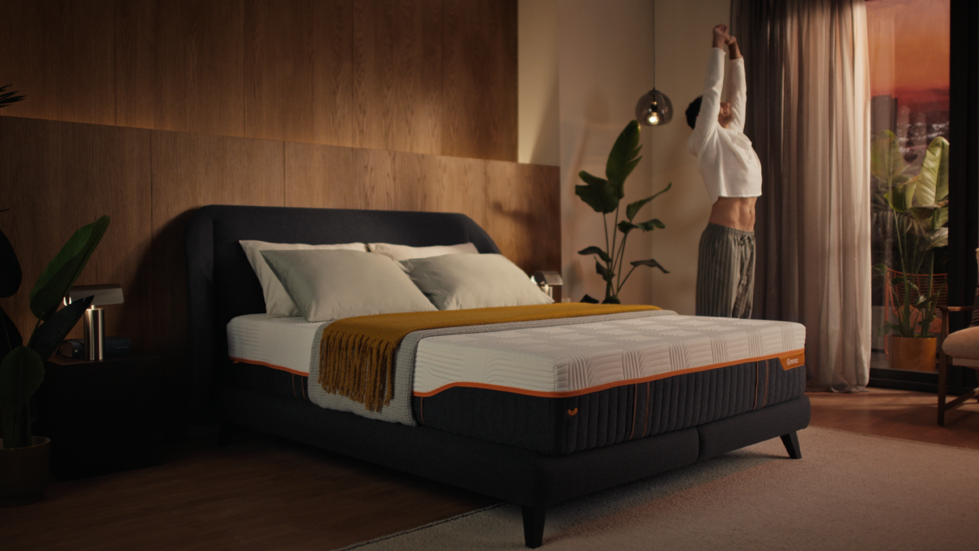 Person stretching after waking next to Emma Performance boxspringbed in a warmly lit, styled bedroom.