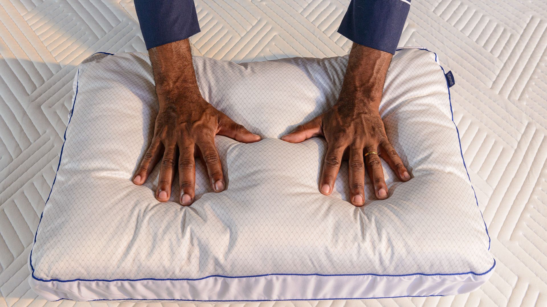 Mãos pressionando um travesseiro branco, travesseiro Antiestresse Emma, apoiado sobre um colchão. A imagem destaca a textura e a maciez do travesseiro.