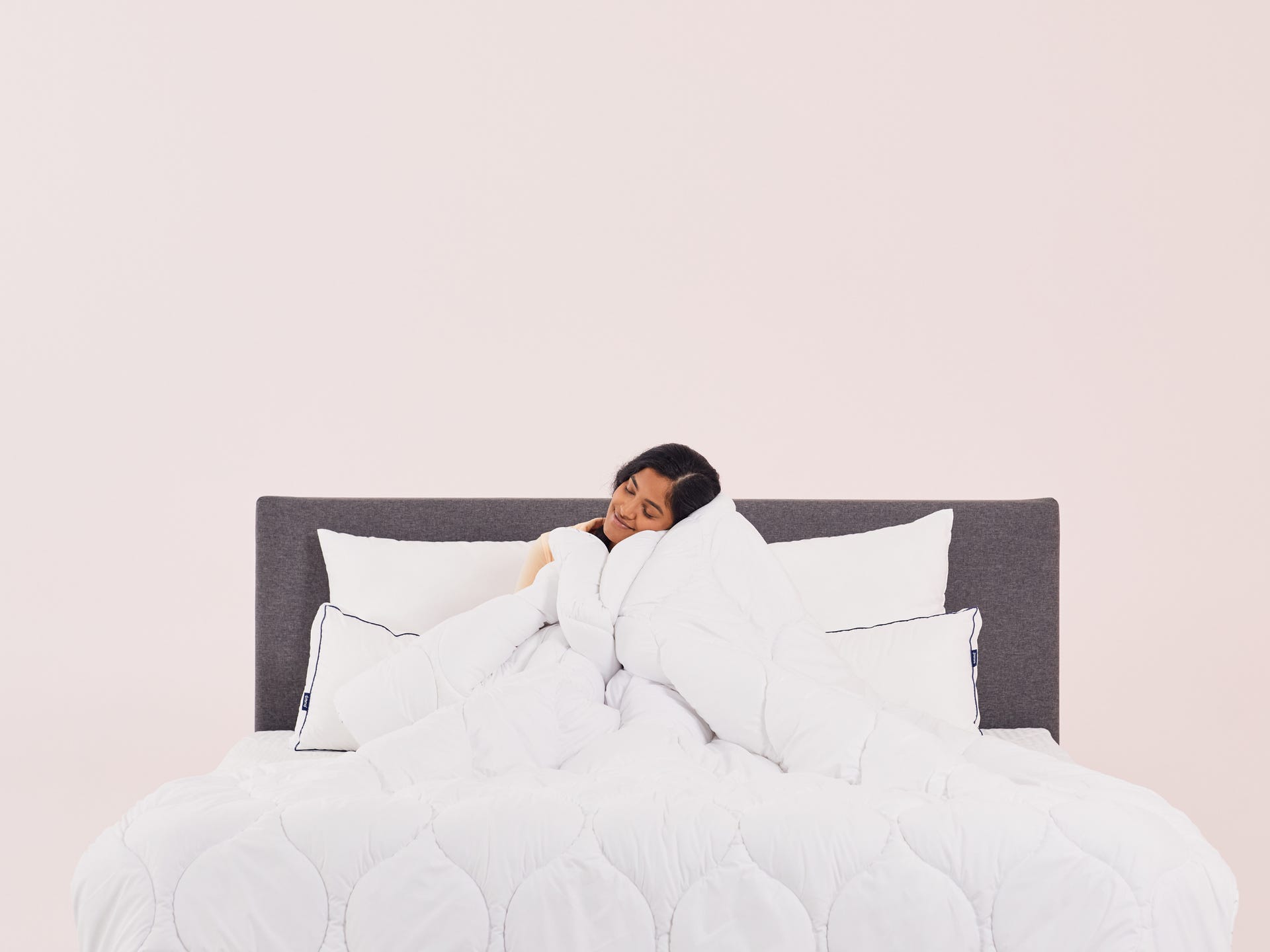 Bed met grijs hoofdbord en wit beddengoed, inclusief twee witte kussens met zwarte bies; bed is netjes opgemaakt en oogt zacht en comfortabel; achtergrond bestaat uit een effen lichtroze muur.