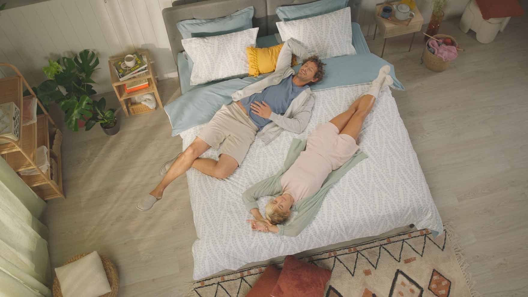 Vue de dessus d’un homme et d’une femme allongés de manière décontractée sur un lit avec une couette blanche et des draps gris clair.