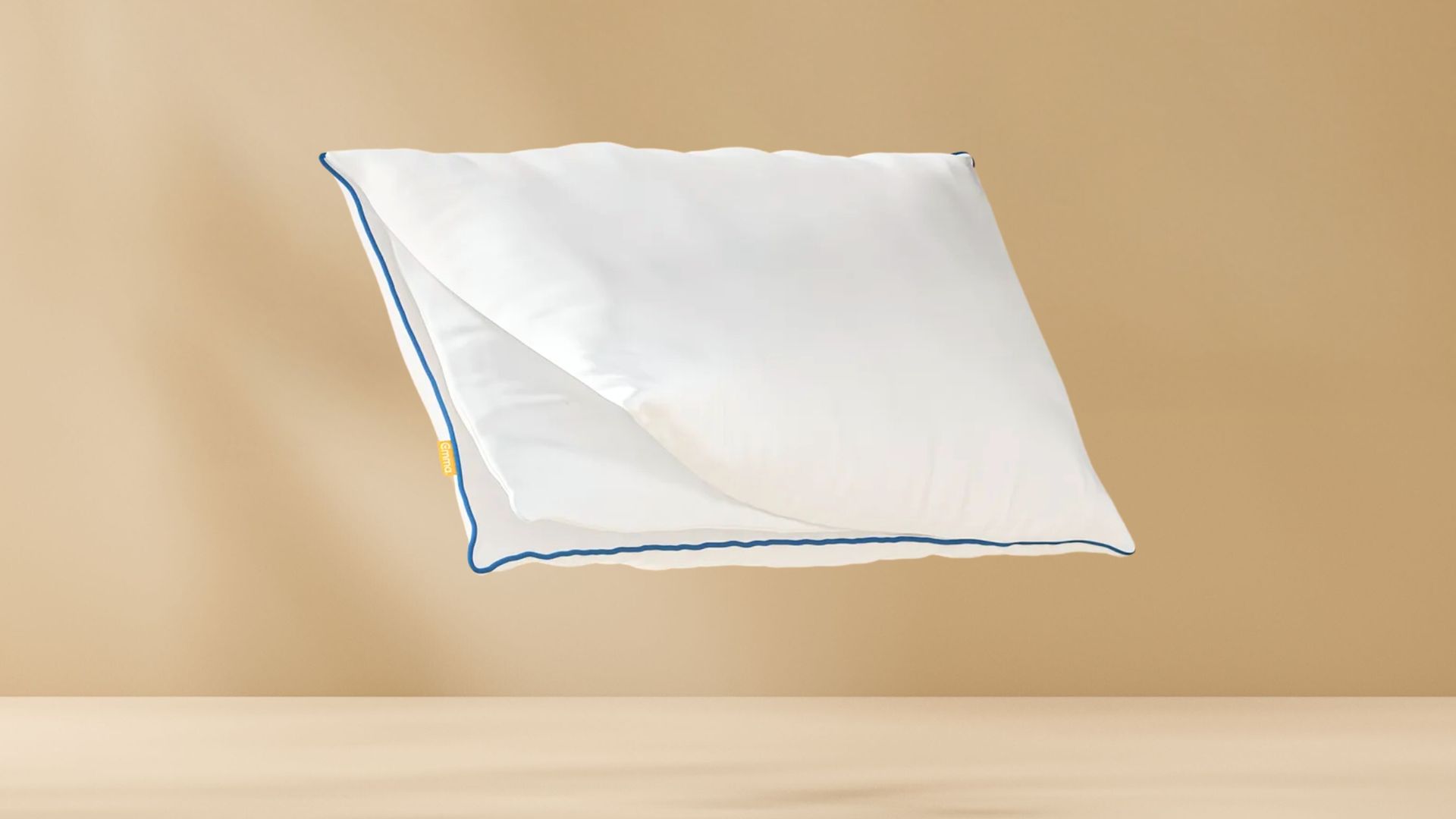 Original Comfort Kussen met witte hoes en blauwe bies zwevend tegen een beige achtergrond, met zichtbaar geel merklabel aan de zijkant.