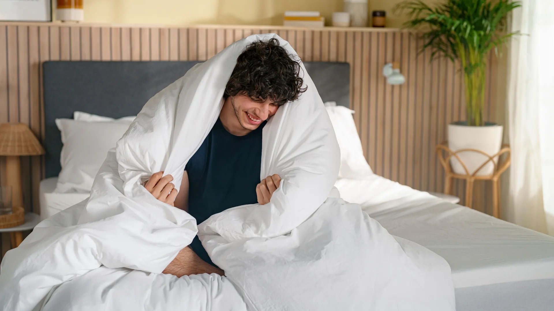 Persoon ligt op bed onder wit Emma dekbed met grijs hoofdbord en witte lakens—lichte slaapkamer met houten vloer, beige gordijnen en decoratieve accenten.