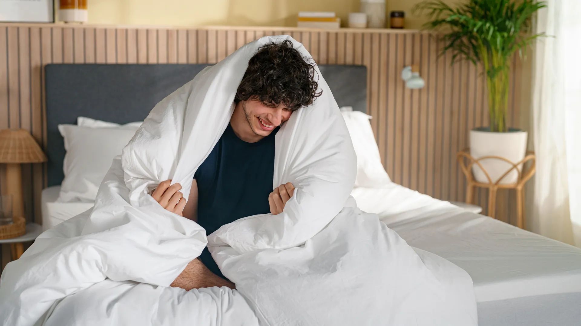 Persoon ligt op bed onder wit Emma dekbed met grijs hoofdbord en witte lakens—lichte slaapkamer met houten vloer, beige gordijnen en decoratieve accenten.