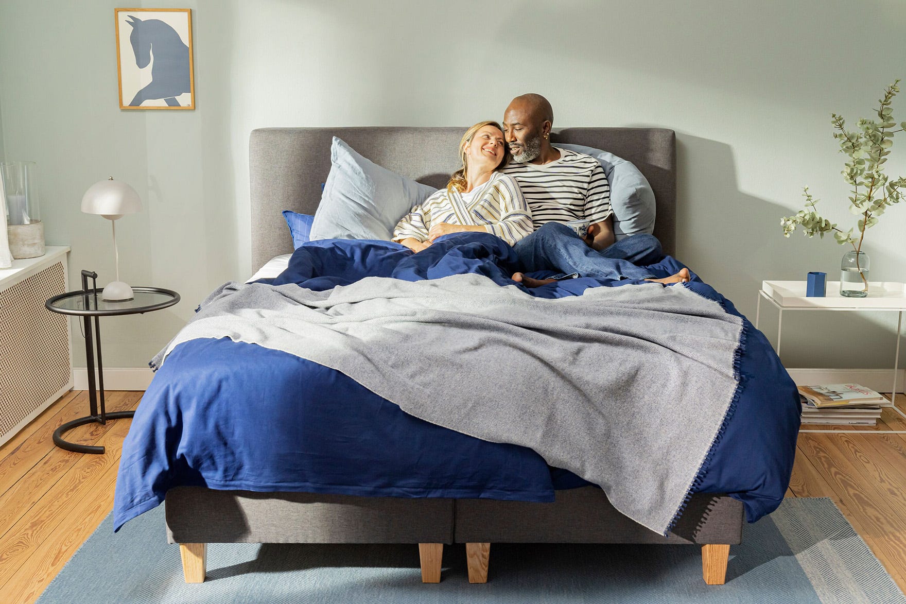 Paar sitzt lachend auf Emma Original Boxspringbett mit grauem Bezug.
