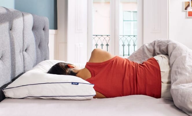 Persoon ligt op zijn zij met hoofd op wit Emma Wolkenkussen, bed met grijs hoofdbord en natuurlijke lichtinval op de achtergrond.