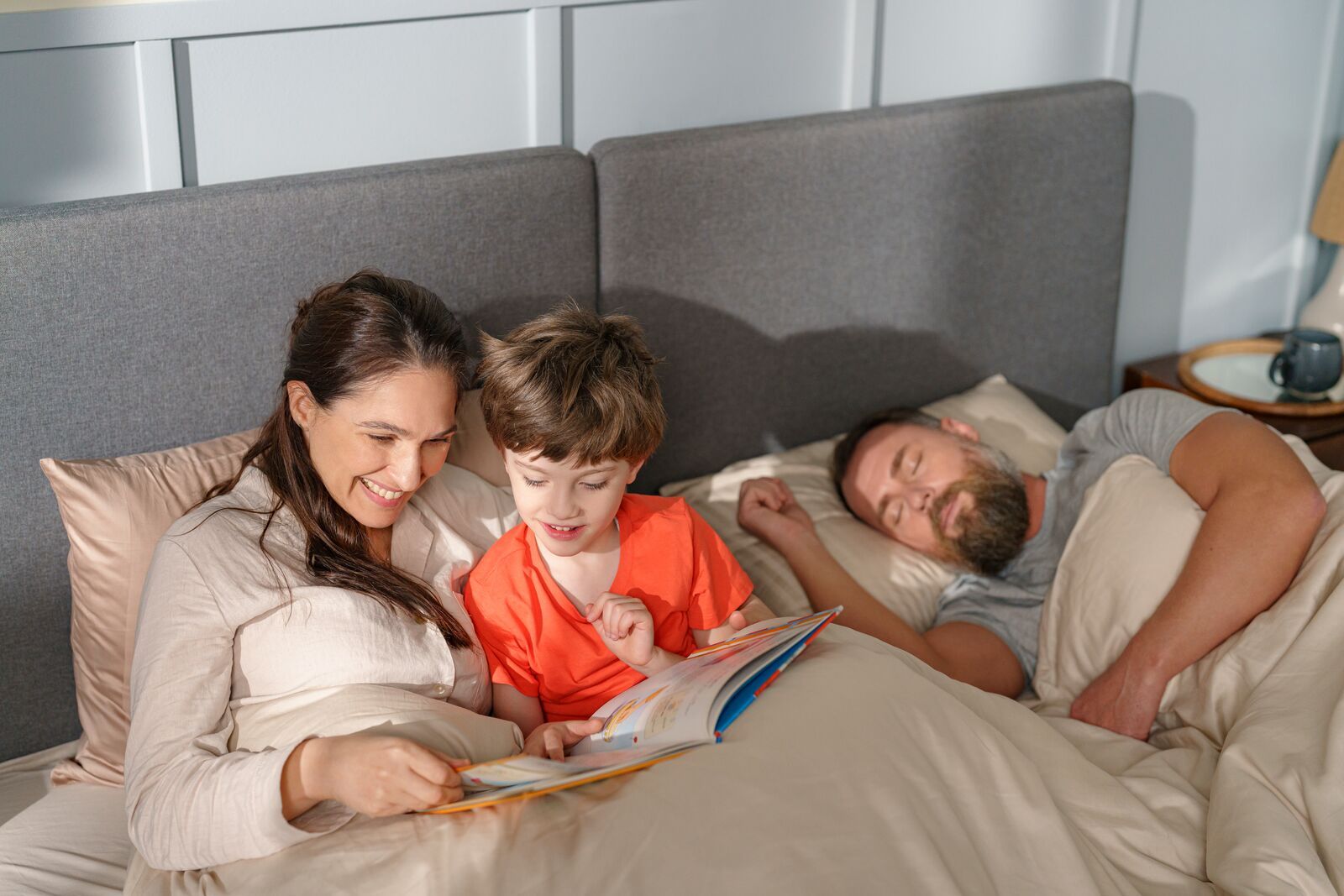 Emma Original Bed met grijs hoofdbord en wit beddengoed—drie personen in bed, één leest een boek aan kind terwijl ander slaapt; nachtkastje met lamp en kopje in lichte slaapkamer.