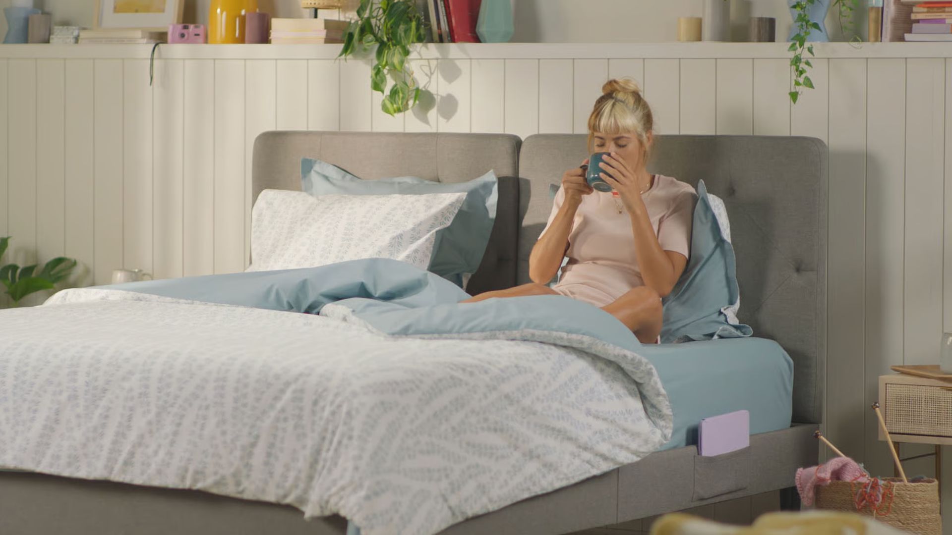 Emma Original Bed met grijs hoofdbord en wit beddengoed—drie personen in bed, één leest een boek aan kind terwijl ander slaapt; nachtkastje met lamp en kopje in lichte slaapkamer.