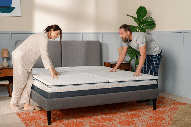 Man en vrouw maken samen een tweepersoonsbed in een moderne slaapkamer met grijs hoofdeinde, nachtkastje, kamerplant en oranje patroonvloerkleed