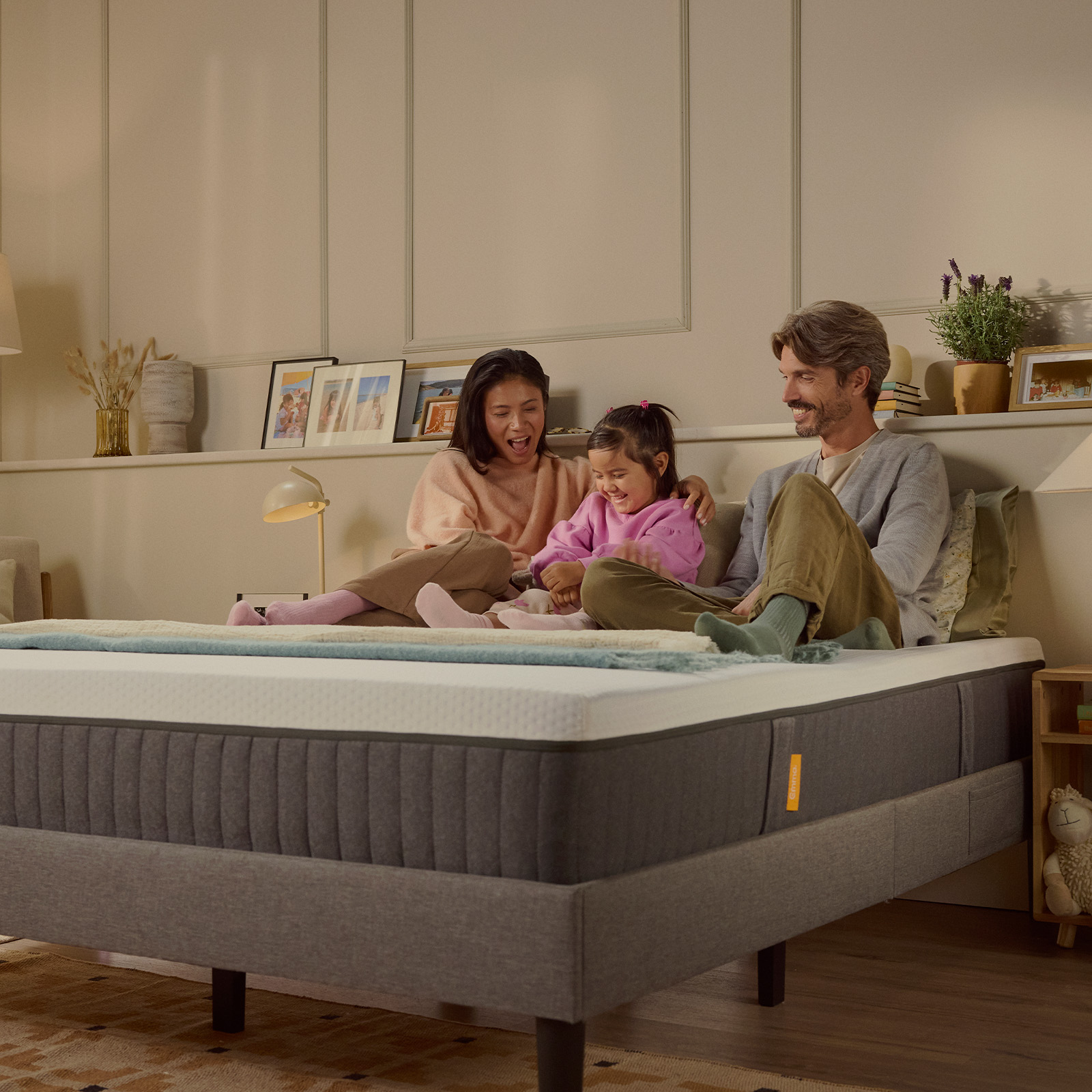 Família de três a rir juntos sobre um colchão Emma Original num quarto com iluminação acolhedora.