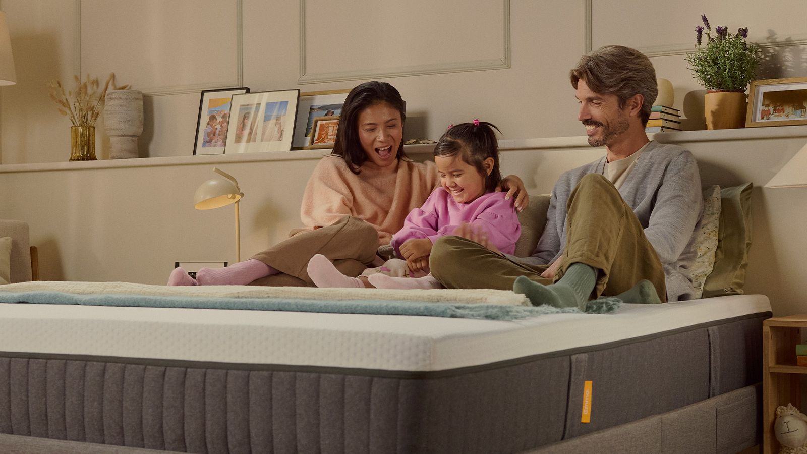 Família de três a rir juntos sobre um colchão Emma Original num quarto com iluminação acolhedora.