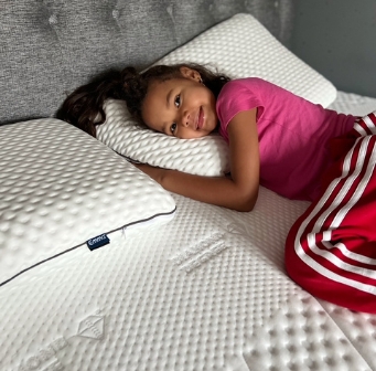 Kind ligt ontspannen op een bed met de Emma Diamond Topper, omringd door witte kussens en een gestoffeerd grijs hoofdbord—een topper met diamantstructuur die zacht en temperatuurregulerend comfort biedt.