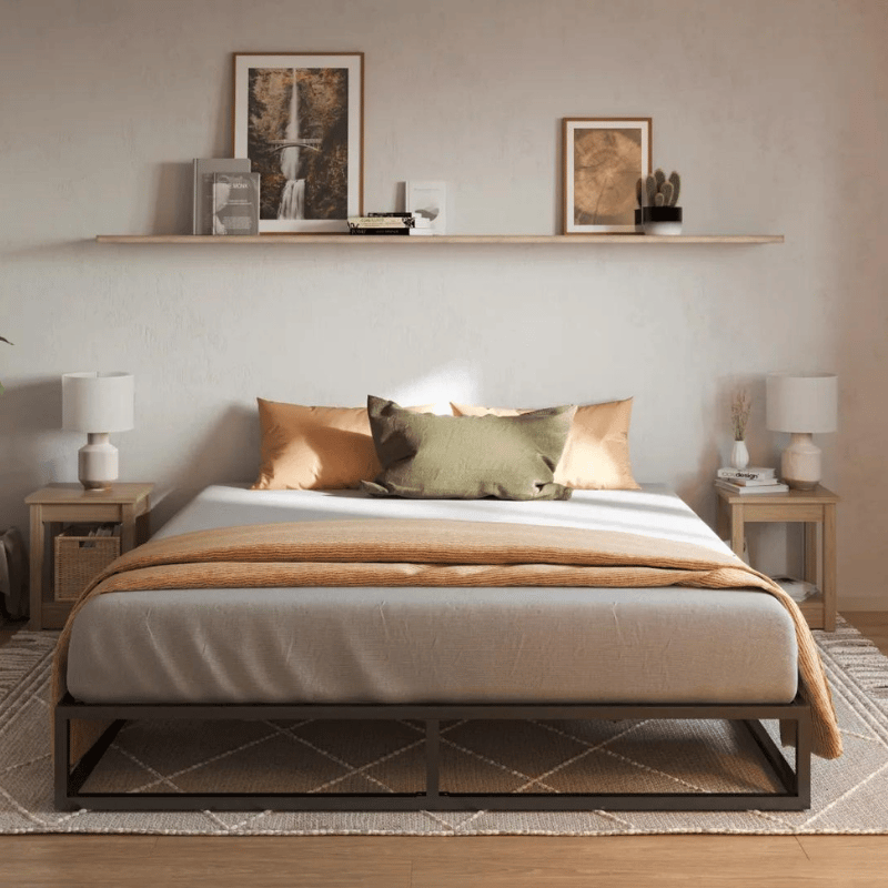 Chambre confortable avec cadre de lit en métal, couette beige et coussins orange sur un sol en bois, avec une étagère exposant des photos encadrées et des objets décoratifs.